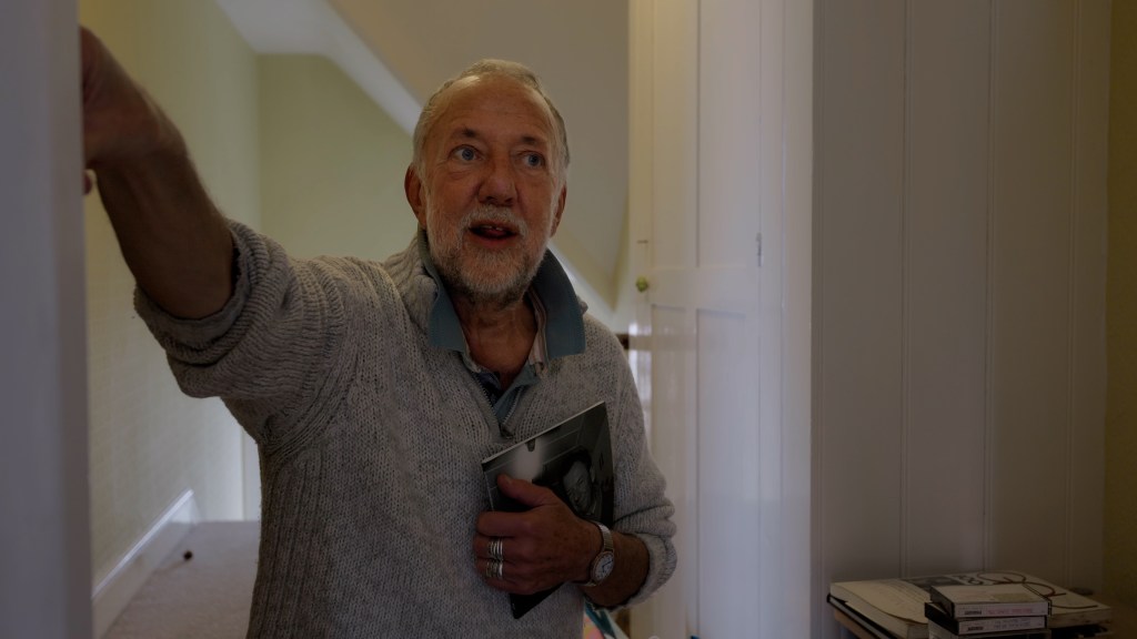A man wearing a wooden jumper holding a book talking to camera